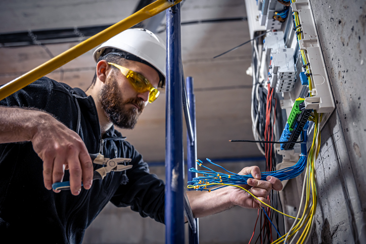 A male electrician works in a switchboard with an electrical connecting cable, connects the equipment with tools