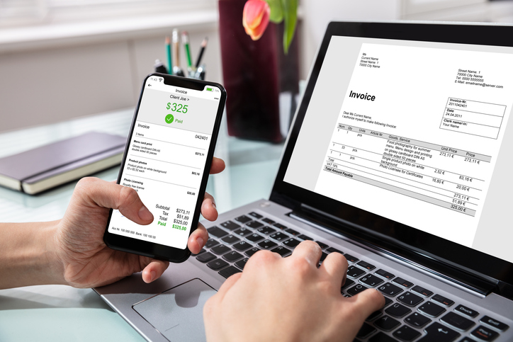 Close-up Of Businessperson Using Laptop While Paying Invoice On Mobilephone In Office