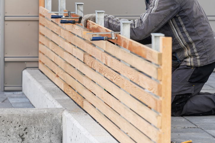 Cropped photo of adult handyman or workman repairing new wooden fence near house using c-clamp